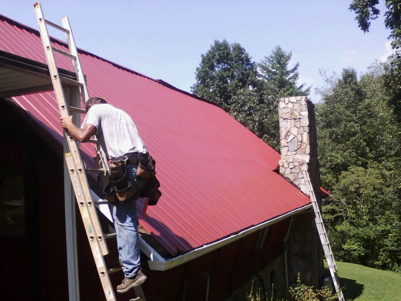 Roofer on a ladder beside a red metal roof and stone chimney — Soffit & Fascia Repair by Roofing Brothers in Shelby